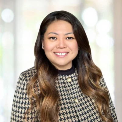A woman with long wavy brown hair wearing a houndstooth patterned jacket smiles at the camera against a softly blurred light background.