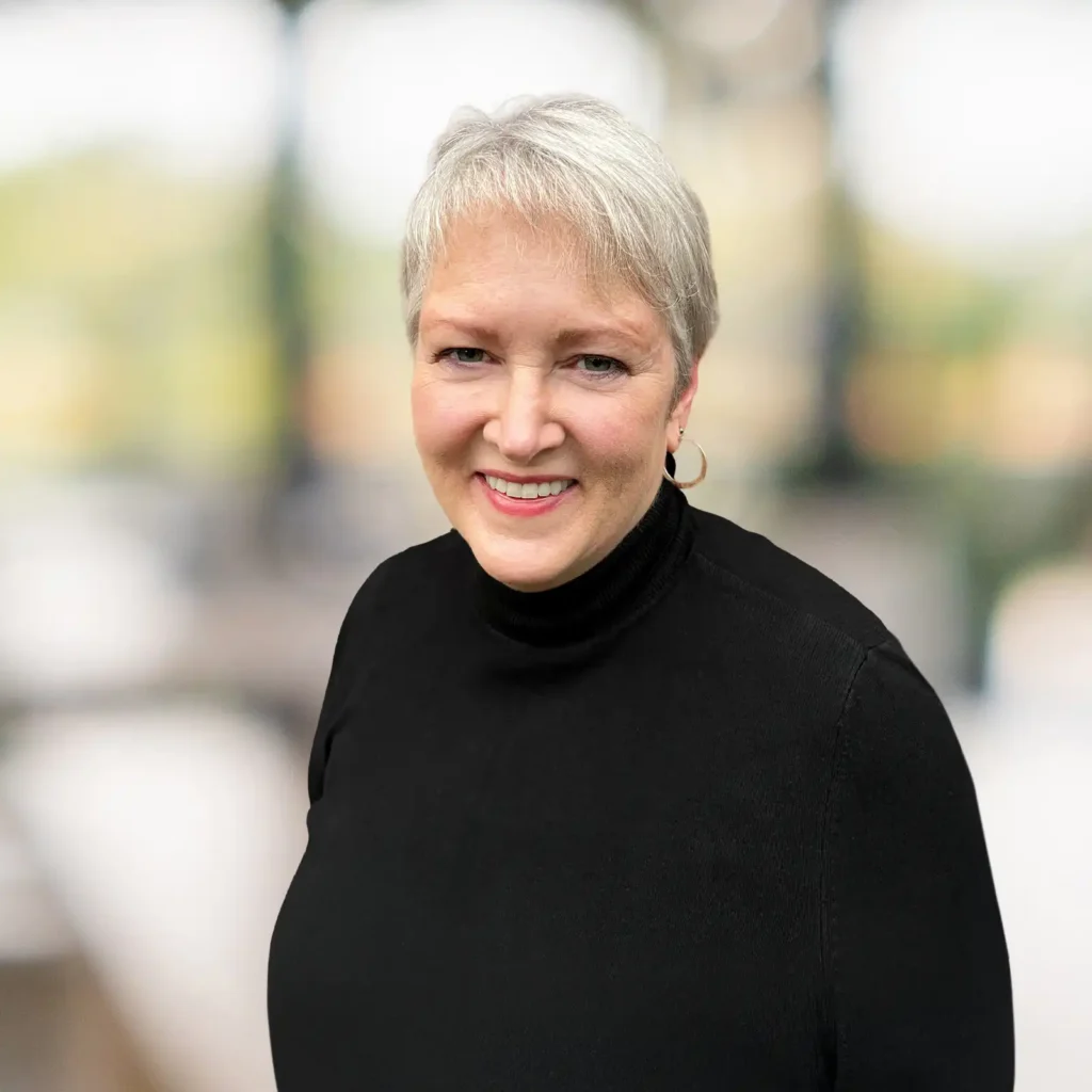 A woman with short gray hair, wearing a black turtleneck and hoop earrings, smiles at the camera. The background is blurred with natural light.