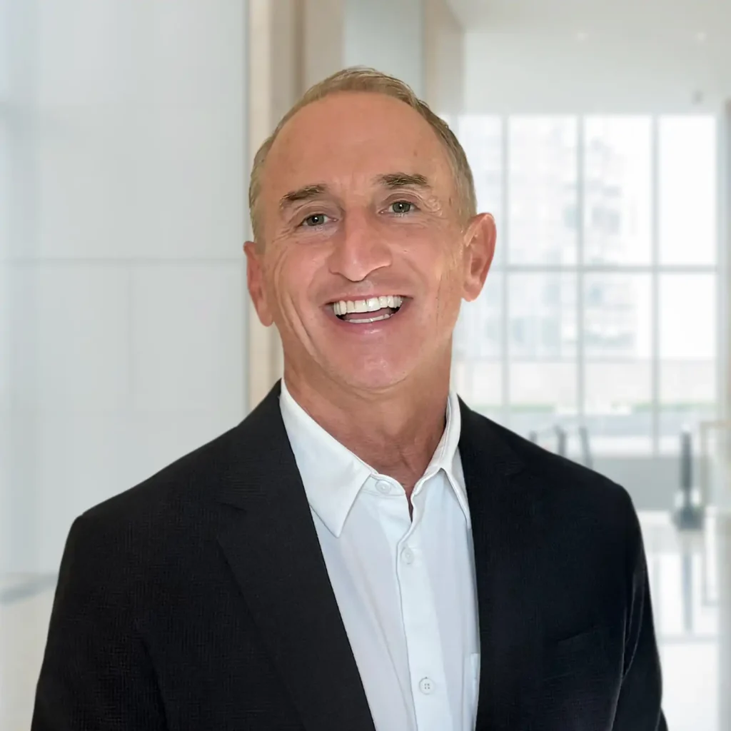 A smiling man in a dark suit jacket and white shirt stands in a bright, modern indoor space with large windows in the background.