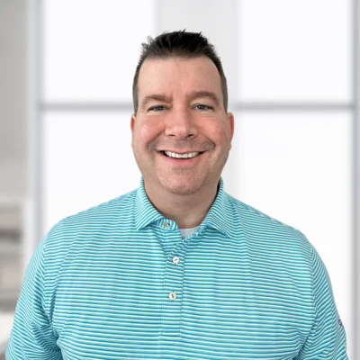 A man wearing a teal and white striped polo shirt smiles at the camera in a bright, modern indoor setting.