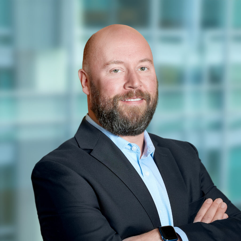 A man with a beard and bald head wearing a suit jacket and light blue shirt stands with arms crossed in front of a blurred office background.