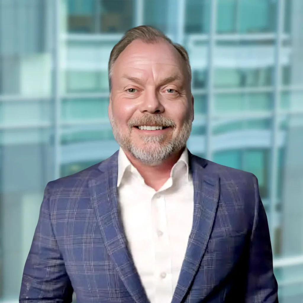 A man with a beard, wearing a blue plaid suit jacket and white shirt, stands smiling in front of a modern glass building background.