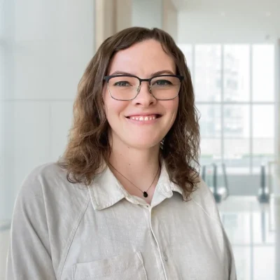A woman with glasses and wavy brown hair smiles at the camera, standing in a bright modern office with large windows in the background.
