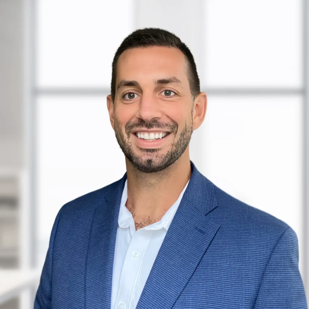 A man with short dark hair and a beard, wearing a blue blazer and white shirt, smiles in front of a blurred office background.