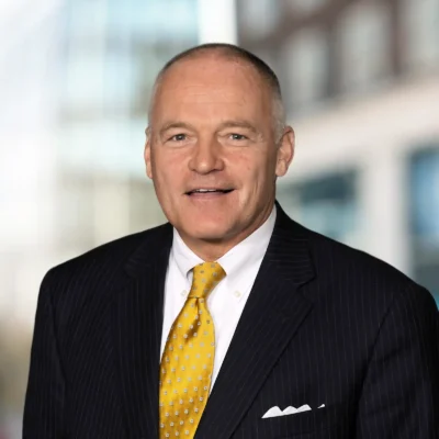A middle-aged man in a dark pinstripe suit, white shirt, and yellow patterned tie stands in front of a blurred office background.
