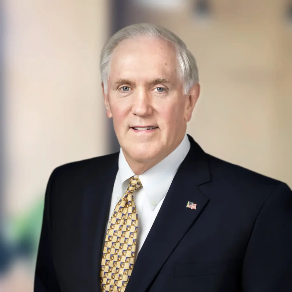 An older man in a dark suit and patterned tie poses for a formal portrait against a blurred neutral background.