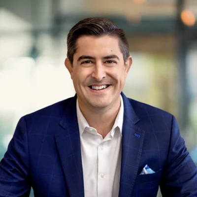 A man in a blue suit and white shirt smiles at the camera; the background is blurred, suggesting an indoor office setting.
