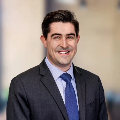 A man in a dark suit, light blue shirt, and blue tie smiles while standing in front of a blurred indoor background.