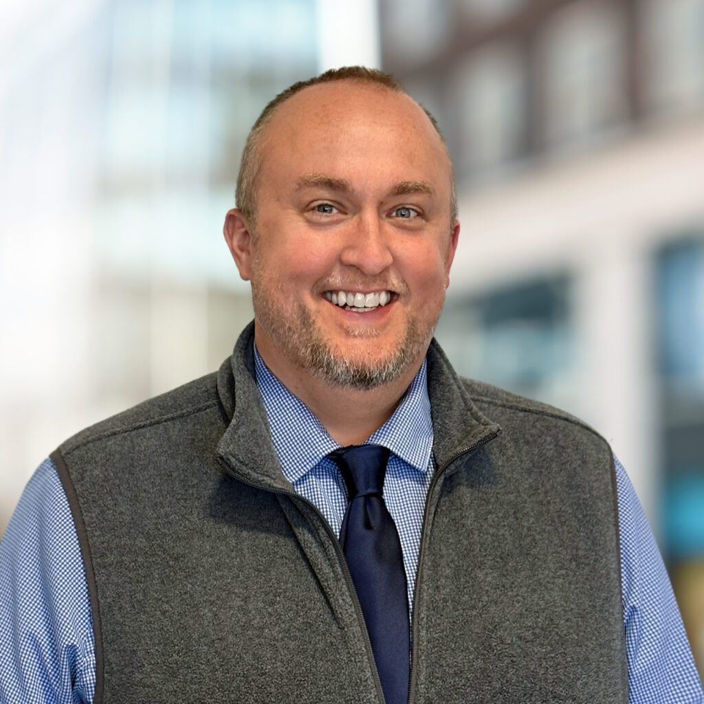 A man with short hair and a trimmed beard smiles at the camera, wearing a blue shirt, tie, and gray vest, with a blurred office background.