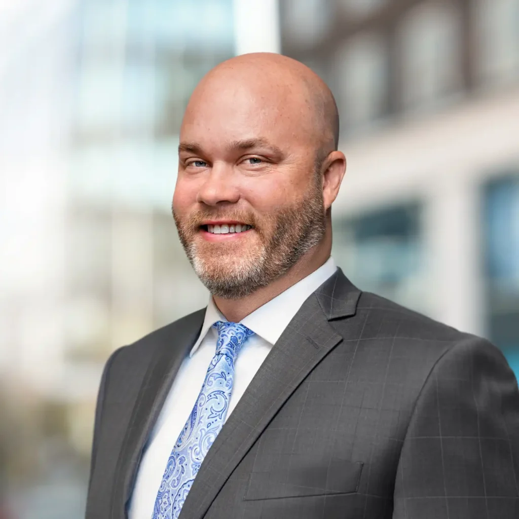 A man with a shaved head and trimmed beard wearing a gray suit, white shirt, and patterned blue tie, standing outdoors with blurred buildings in the background.