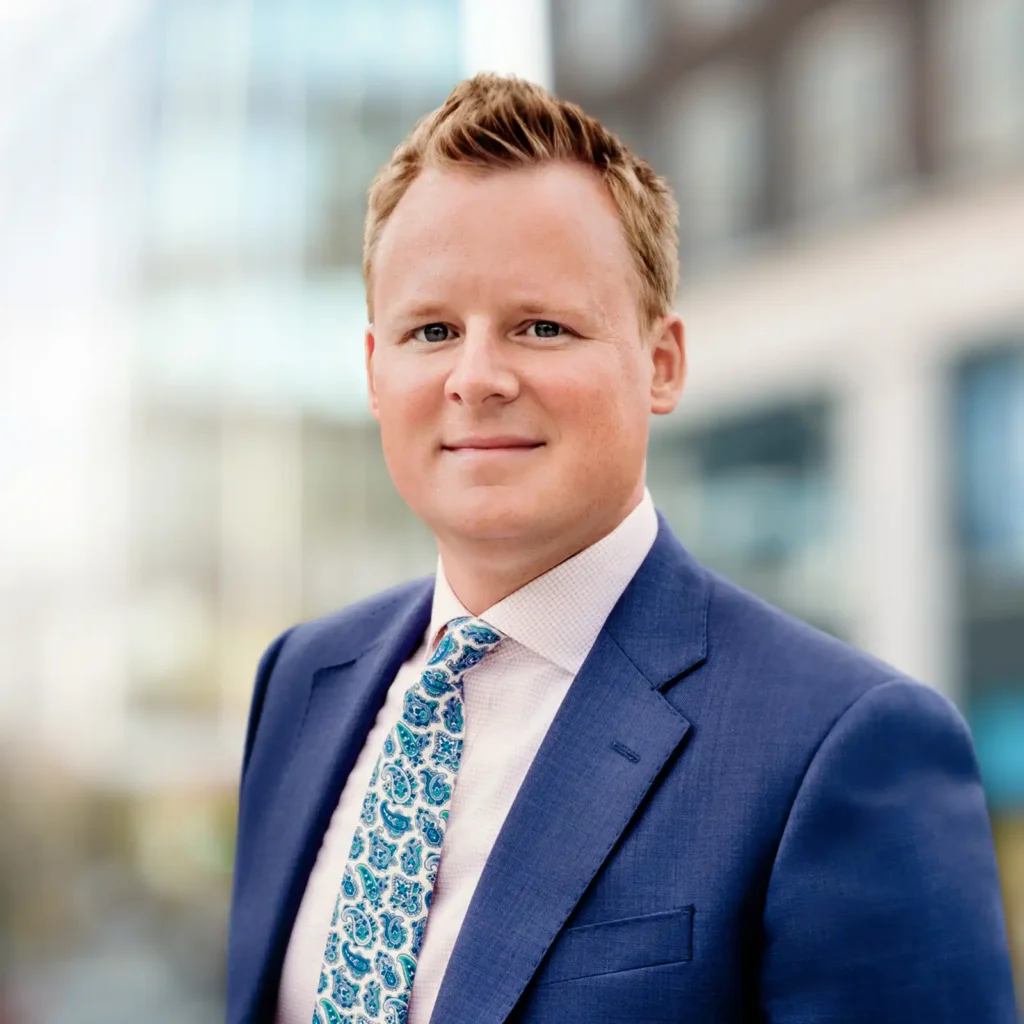 A man in a blue suit and patterned tie stands outdoors in an urban setting with blurred buildings in the background.