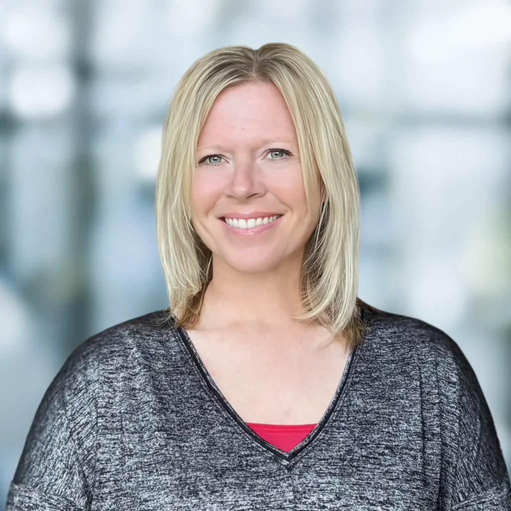 A woman with straight blonde hair, wearing a grey top over a red shirt, smiles at the camera against a blurred indoor background.