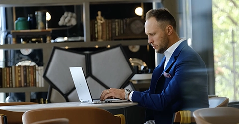A man in a blue blazer sits at a round table working on a laptop in a modern, book-lined room.