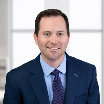 A man in a blue suit and tie smiles at the camera in an office setting with large windows in the background.