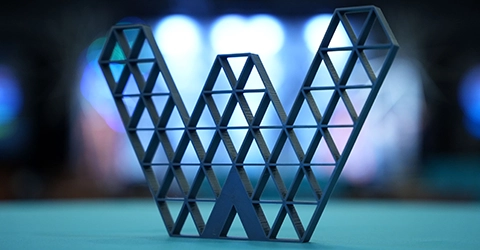 A metallic sculpture shaped like the letter "W" featuring a triangular lattice design, displayed against a blurred background.