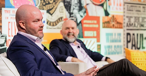 Two men in suits sit and speak on a panel in front of a colorful wall covered with various posters and signs.