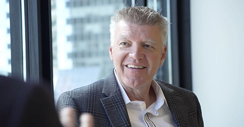 A middle-aged man in a checked blazer and white shirt sits indoors by a window, smiling and engaged in conversation.