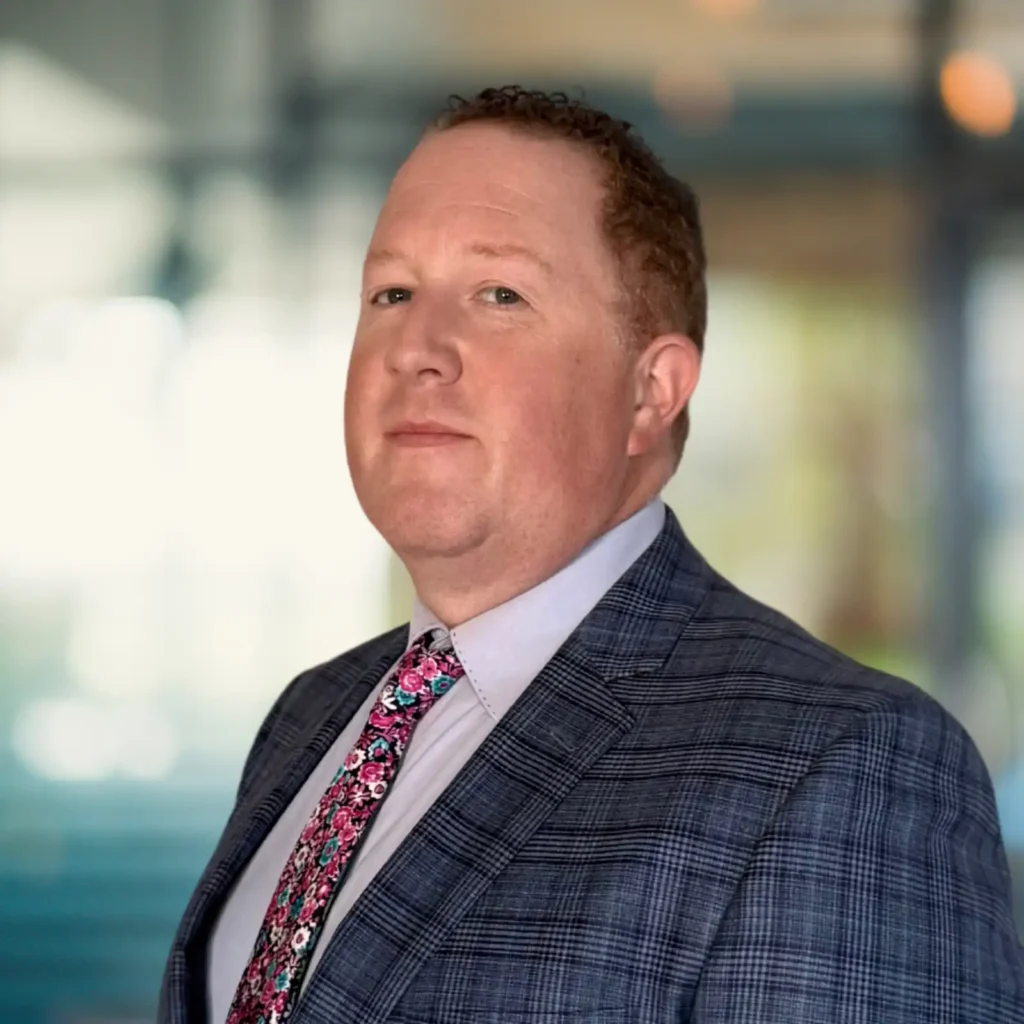 A man with short curly hair in a checked suit jacket, light shirt, and floral tie stands in front of a blurred indoor background.