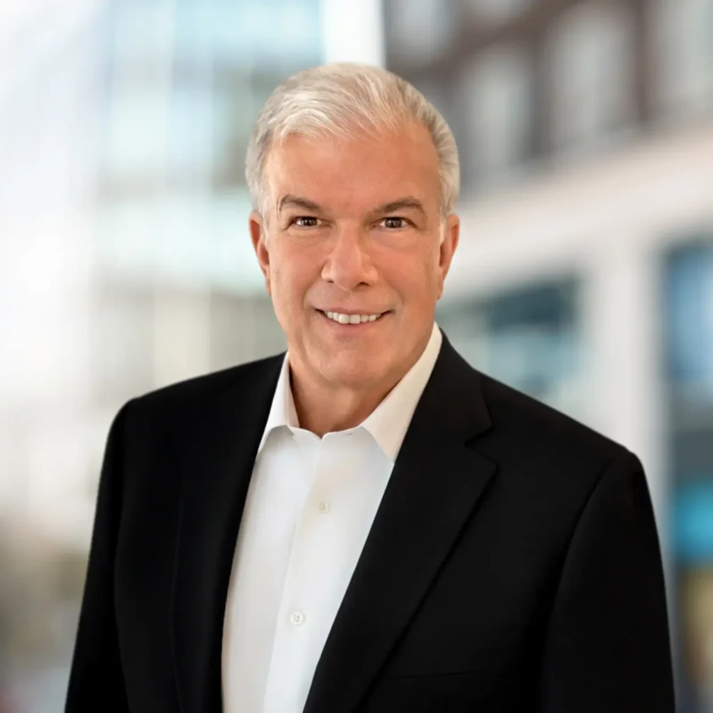 A middle-aged man with gray hair wearing a white shirt and black suit jacket smiles at the camera, with a blurred cityscape in the background.
