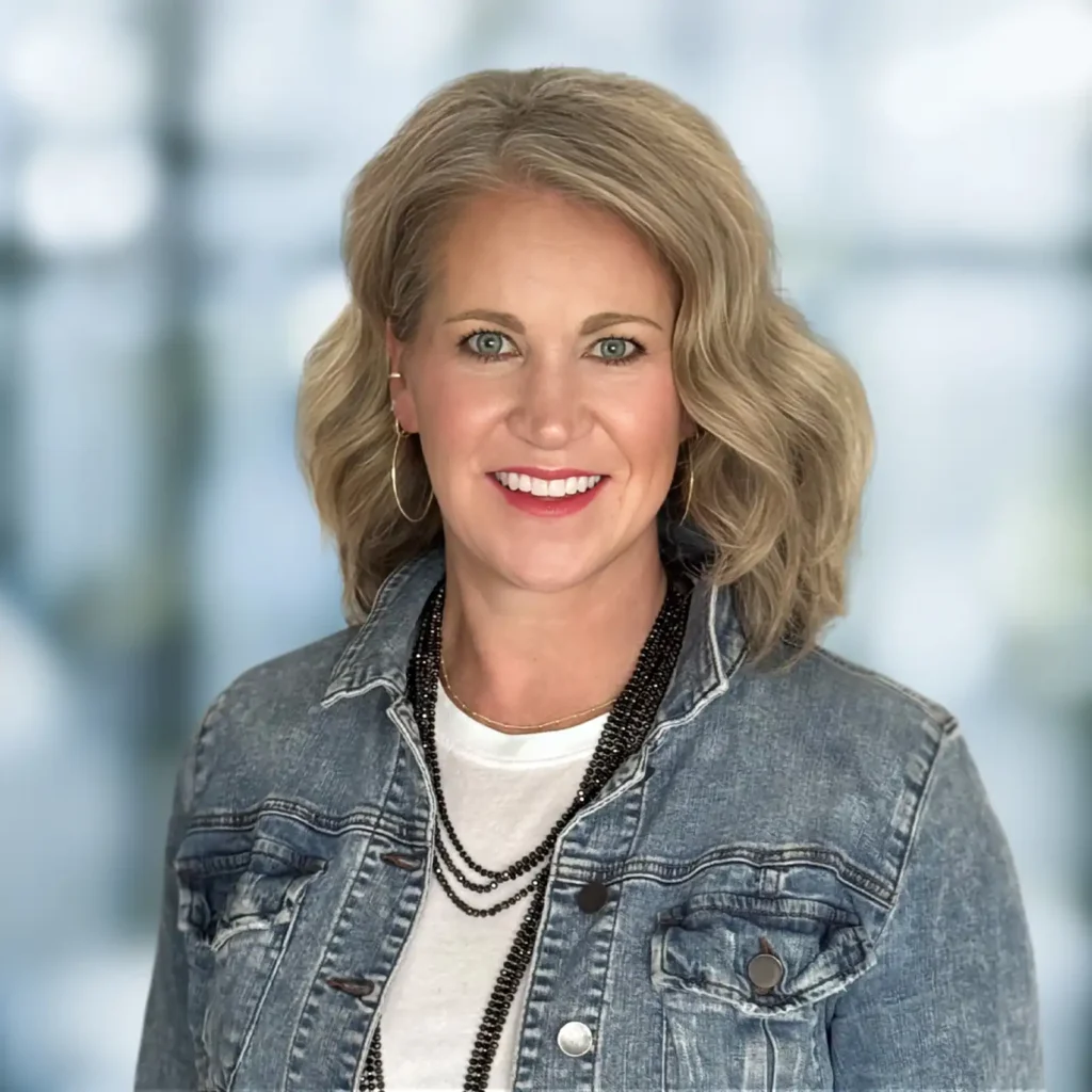 A woman with shoulder-length blonde hair wearing a denim jacket, white shirt, and layered black necklace, smiling at the camera with a blurred background.