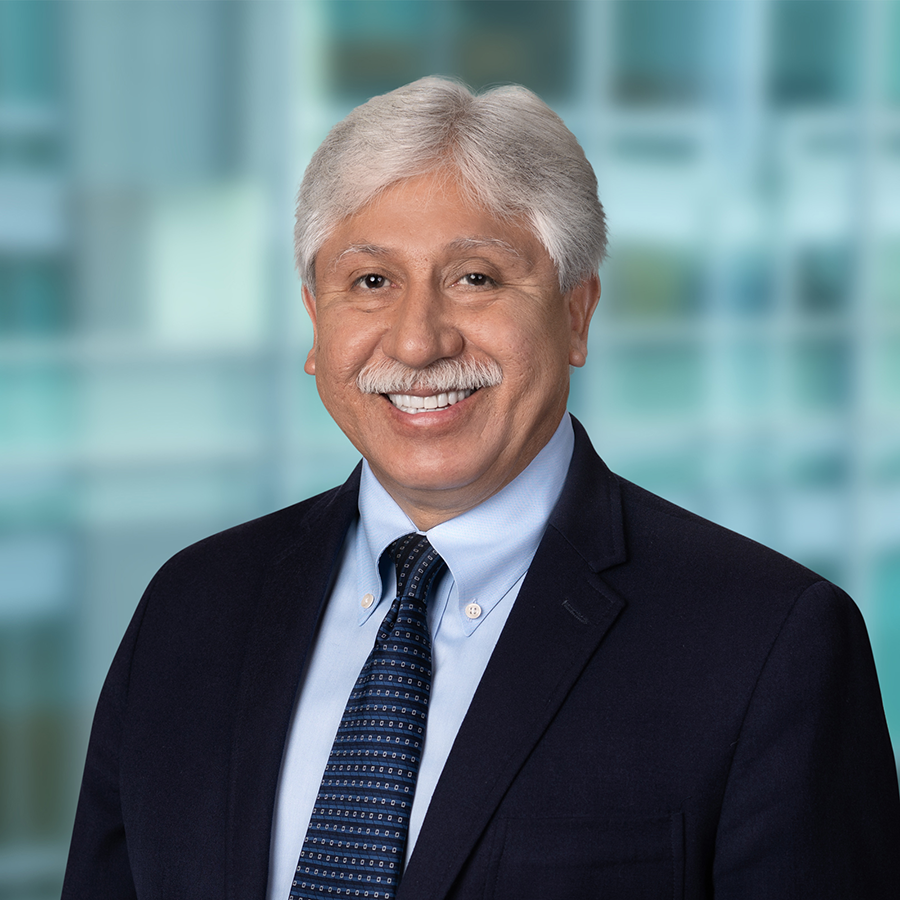 Smiling man with white hair and mustache wearing a dark suit, light blue shirt, and patterned tie, standing in front of a blurred office background.