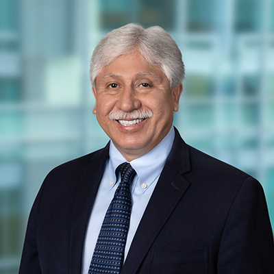 Smiling man with gray hair and mustache wearing a dark suit, light blue shirt, and patterned tie, posed in front of a blurred office background.