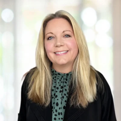 Woman with long blonde hair wearing a green blouse with black spots and a black jacket, smiling, with a blurred light background.