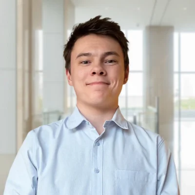 A young man in a light blue button-up shirt stands indoors in a bright, modern office setting, looking directly at the camera and smiling slightly.
