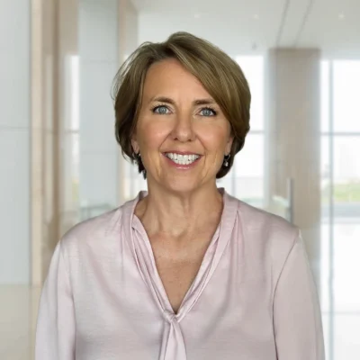 A woman with short light brown hair, wearing a light pink blouse, stands smiling in a modern, bright office space with large windows.