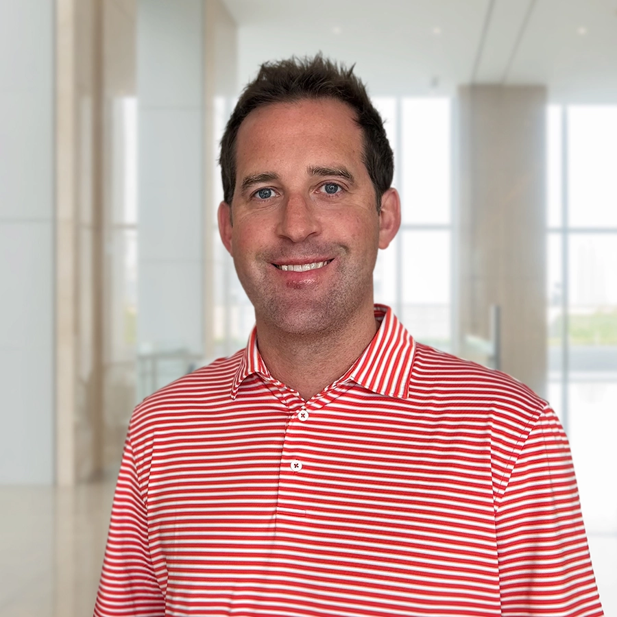 A man wearing a red and white striped polo shirt stands smiling in a bright, modern office lobby with large windows.