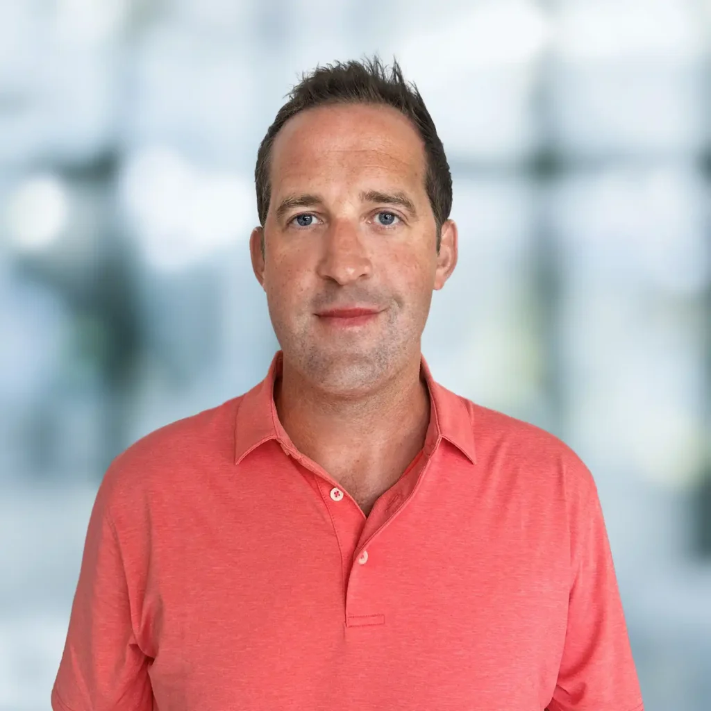 A man wearing a coral polo shirt stands in front of a blurred background with soft natural light.