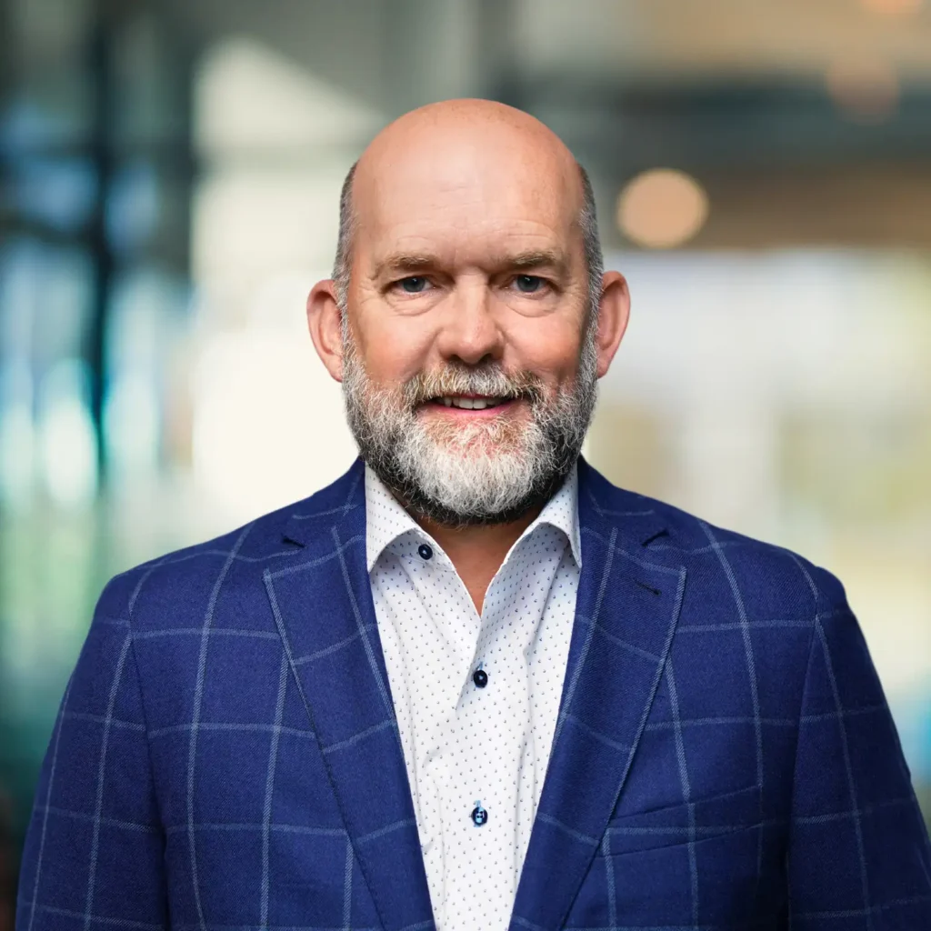 A middle-aged man with a bald head and gray beard wearing a blue plaid blazer and white dress shirt stands indoors with a blurred background.