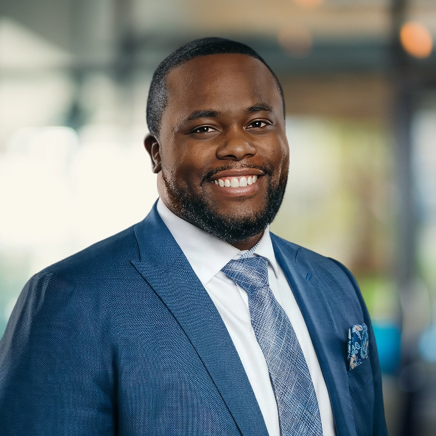 A man in a blue suit and tie smiles at the camera while standing indoors with a blurred background.
