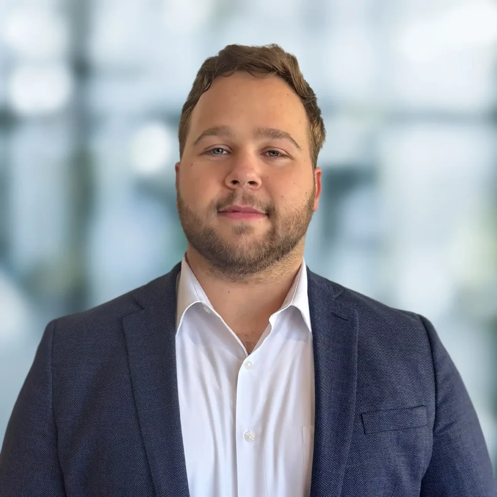 A man with short brown hair and a beard wearing a white shirt and dark blazer stands in front of a blurred indoor background.