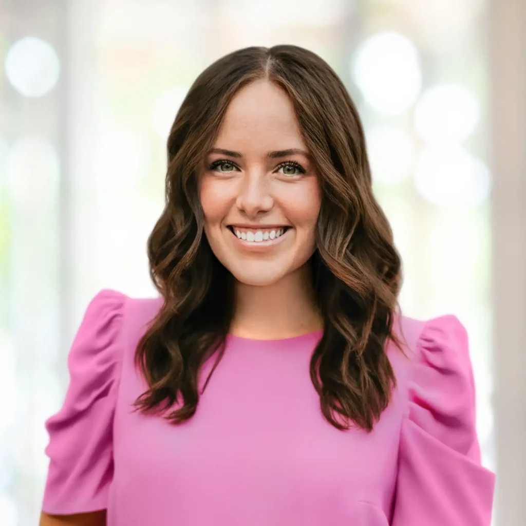 A woman with wavy brown hair wearing a bright pink puff-sleeve top smiles at the camera against a softly blurred background.