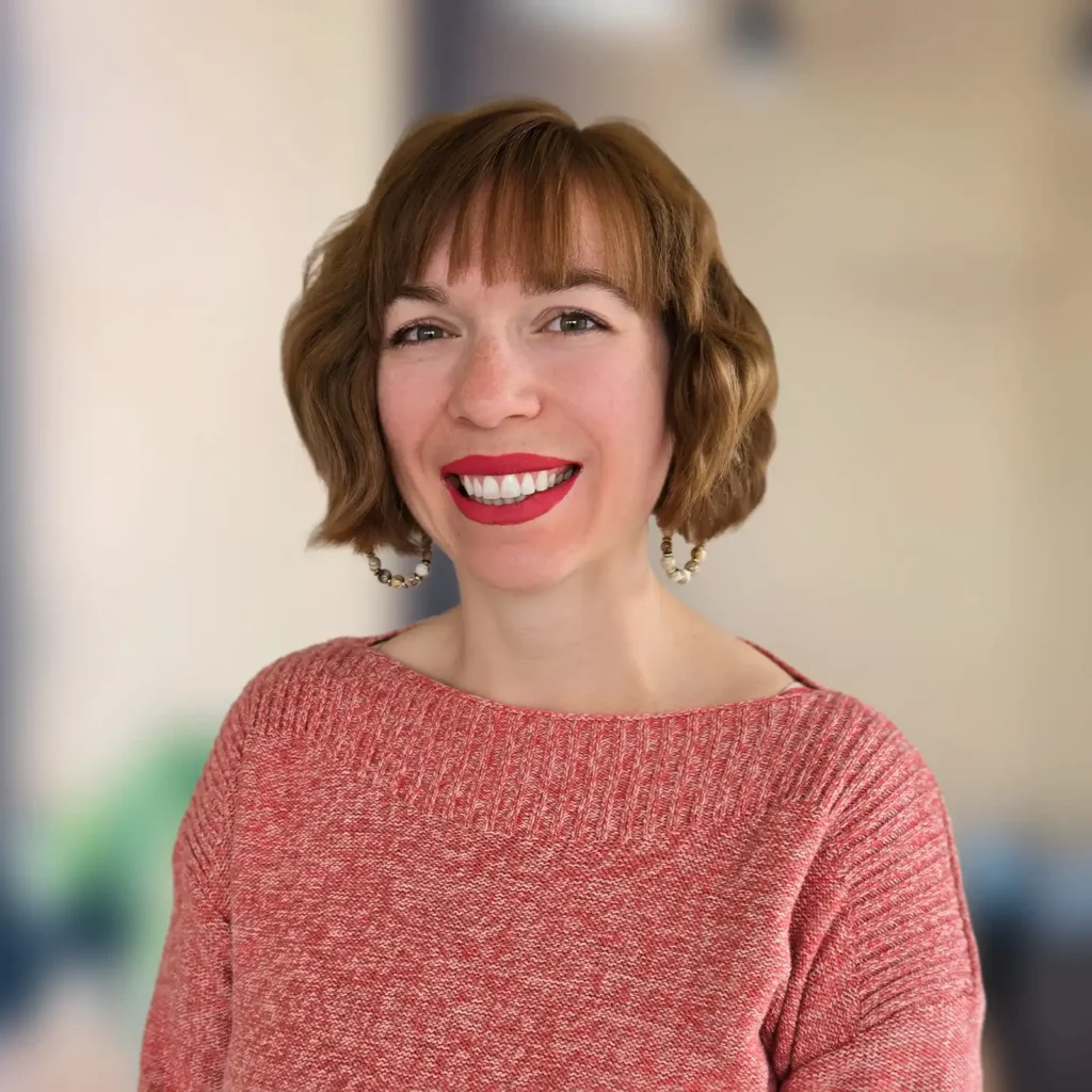 A woman with short brown hair and red lipstick smiles while wearing a red sweater and hoop earrings, standing in front of a blurred indoor background.