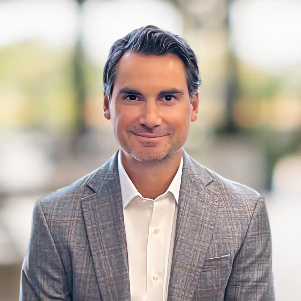 A man wearing a gray plaid suit jacket and white shirt sits indoors, smiling slightly, with a blurred background.