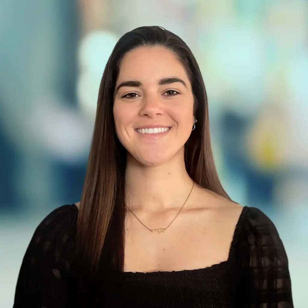 A woman with straight brown hair wearing a black top and a necklace smiles at the camera against a blurred background.