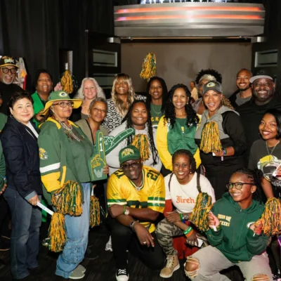 A group of people wearing Green Bay Packers gear pose together indoors, holding pom-poms and foam fingers, with arcade machines in the background.