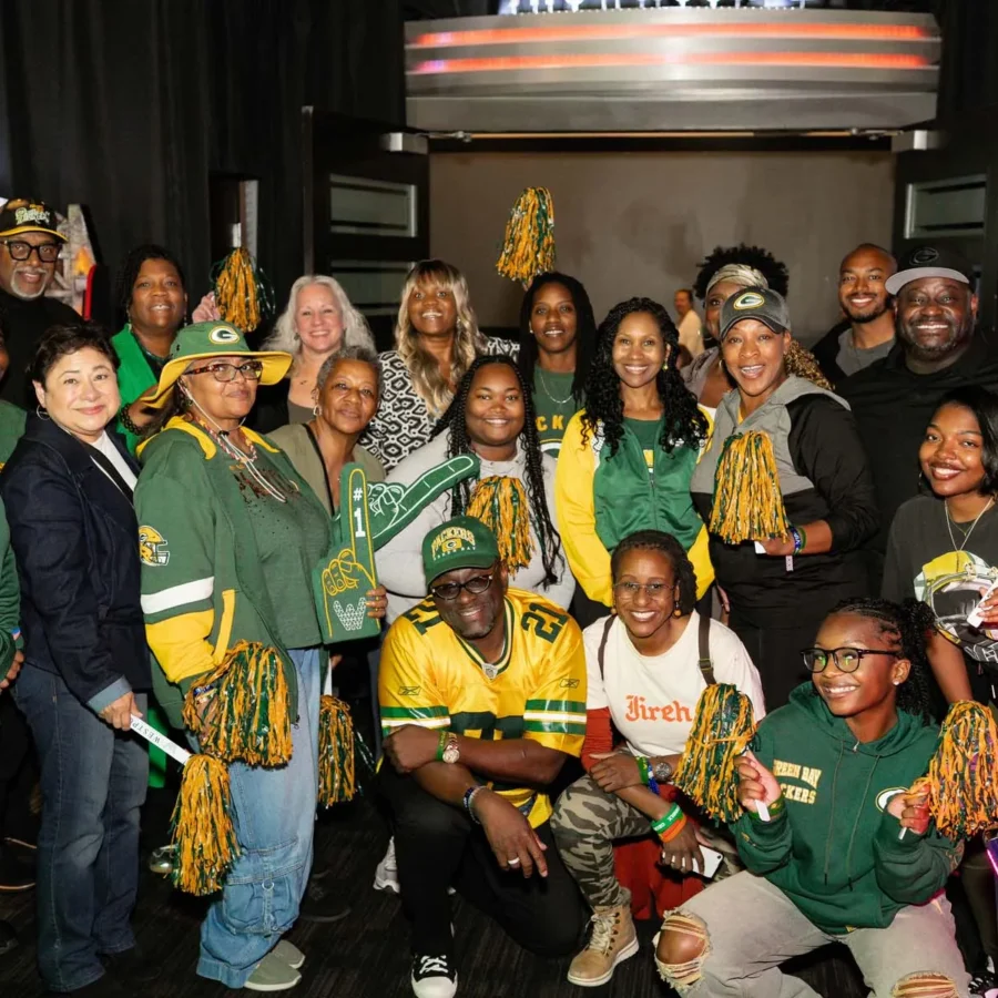A group of people wearing Green Bay Packers gear pose together indoors, holding pom-poms and foam fingers, with arcade machines in the background.