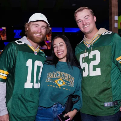Three people wearing Green Bay Packers shirts pose together and smile indoors, with other people and screens visible in the background.