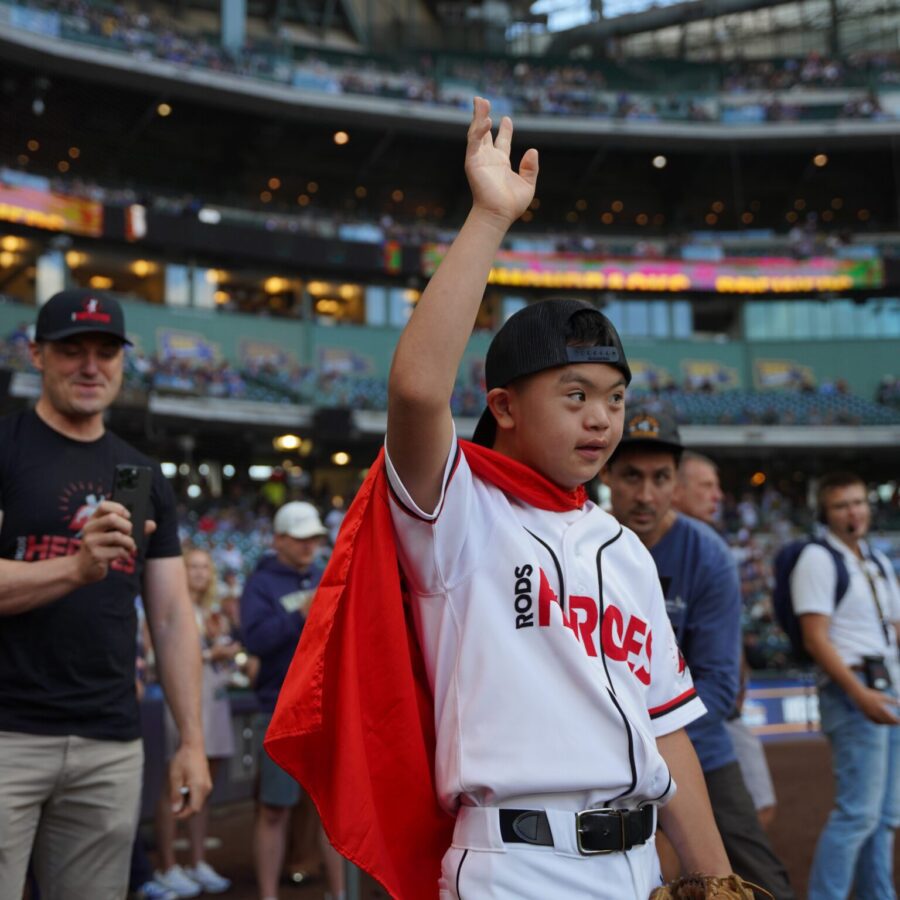 A young baseball player in a white uniform and red cape raises his hand on a baseball field, with spectators and other people in the background.