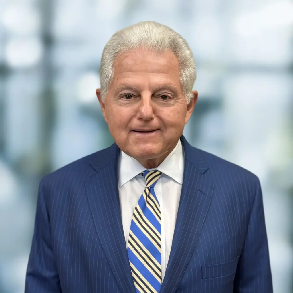 Older man with short gray hair wearing a blue pinstripe suit, white shirt, and striped tie, standing in front of a blurred background.