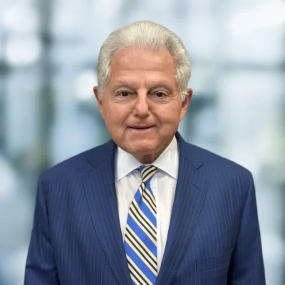 Older man with short gray hair wearing a blue pinstripe suit, white shirt, and striped tie, standing in front of a blurred background.