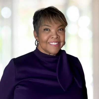 Woman with short hair wearing a dark purple top with a bow stands indoors, smiling at the camera. The background is softly blurred.