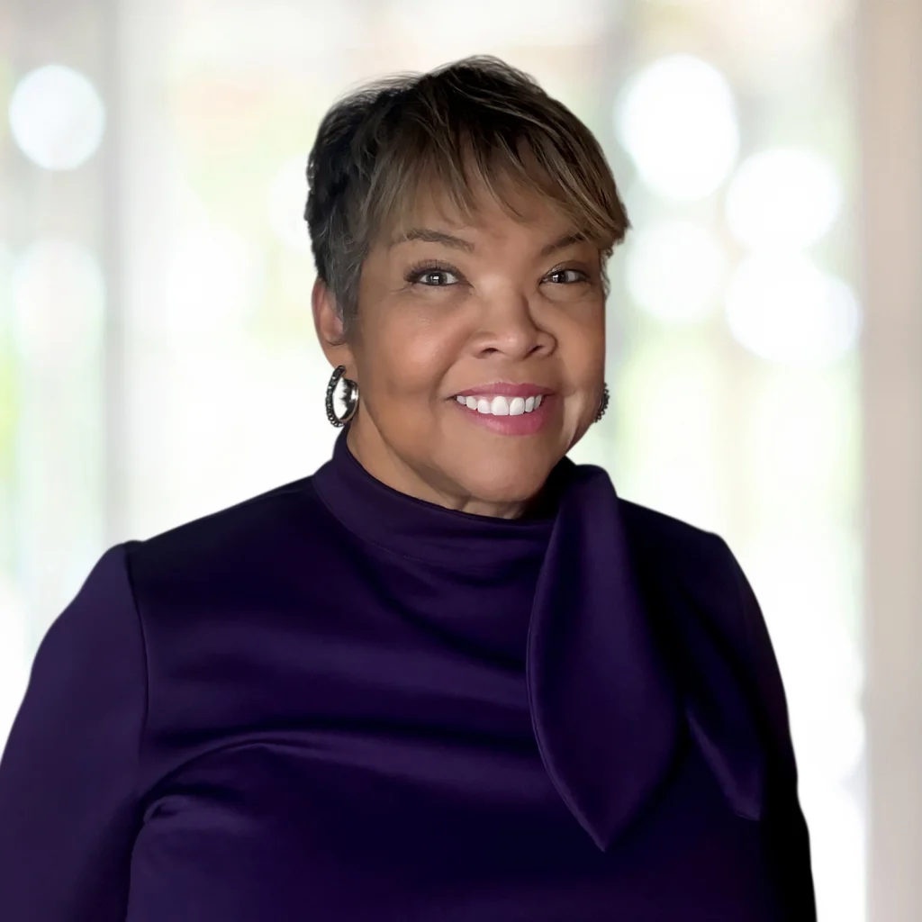 A woman with short hair and hoop earrings, wearing a purple top with a decorative collar, smiles at the camera against a softly blurred background.