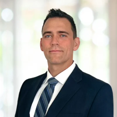 A man wearing a dark suit, white shirt, and striped tie poses in front of a blurred light background.