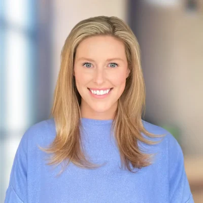 A woman with long blonde hair and a blue top smiles at the camera, standing in a softly blurred indoor setting.
