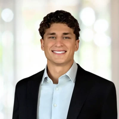 A young man with short curly hair smiles, wearing a light blue shirt and black blazer, standing in front of a blurred light background.
