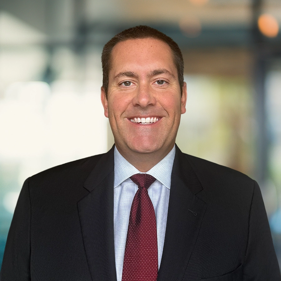 A man in a dark suit, light shirt, and red tie smiles at the camera in a blurred office setting.